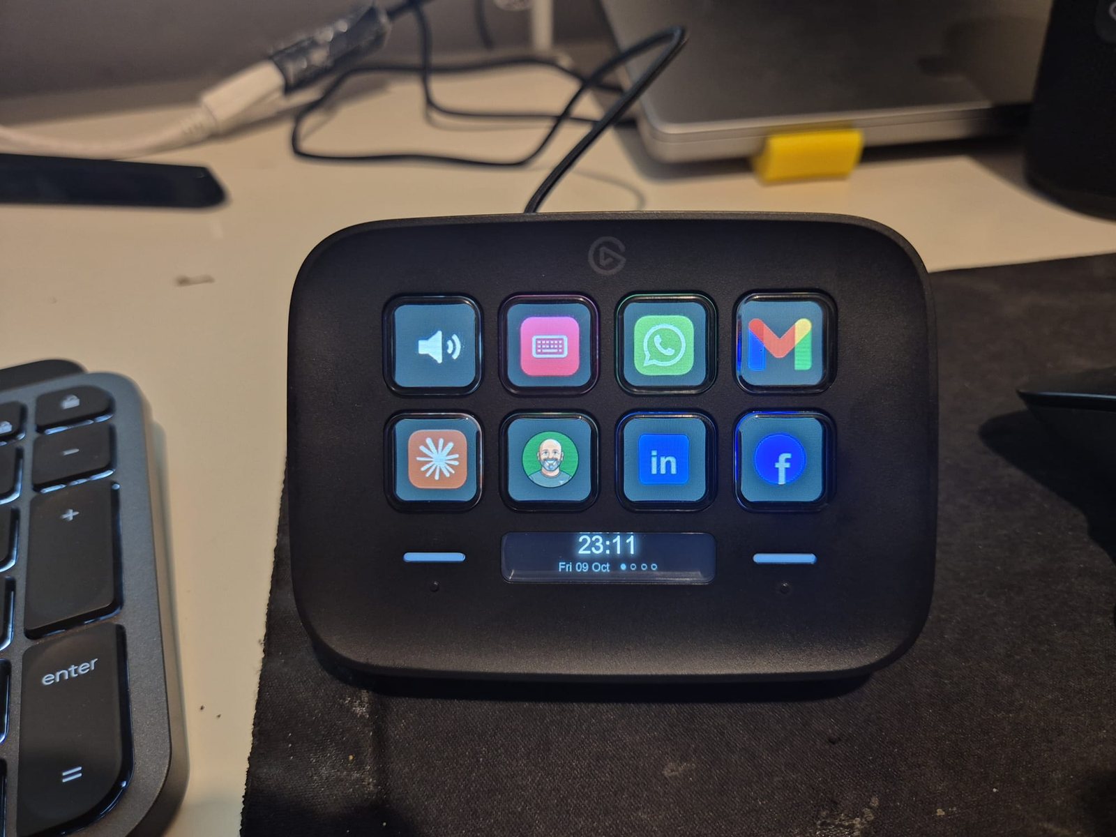 The same Stream Deck at night with icon keys: a speaker, a keyboard layout fix key, WhatsApp, Gmail, Claude, a personal avatar, LinkedIn and Facebook, and a clock with page dots on the display.